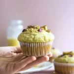 A hand gently holds one pistachio muffin, with other muffins showing in the background.