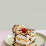 A piece of banana split cake, topped with a cherry, chopped nuts, two slices of banana, and drizzled with chocolate sauce, sits on a green plate.