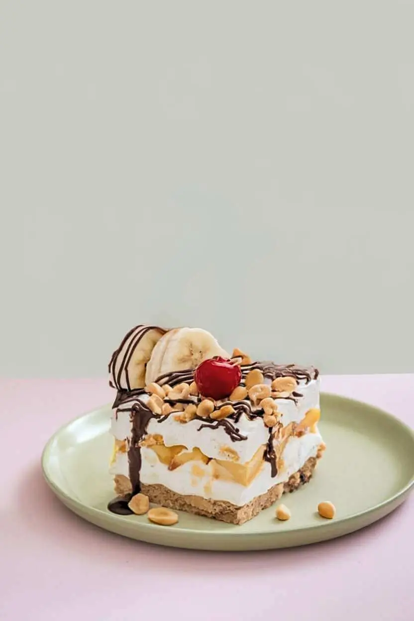 A piece of banana split cake, topped with a cherry, chopped nuts, two slices of banana, and drizzled with chocolate sauce, sits on a green plate.