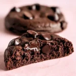 Half of an almond flour chocolate cookie rests on a light pink surface, positioned Infront of another cookie.