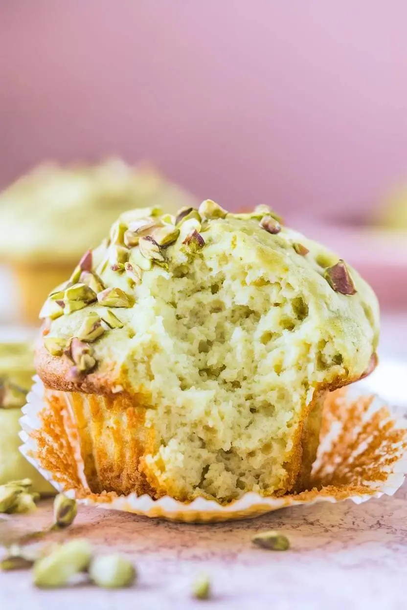 One pistachio muffin topped with chopped pistachios, with a bite taken out, on a light pink surface.