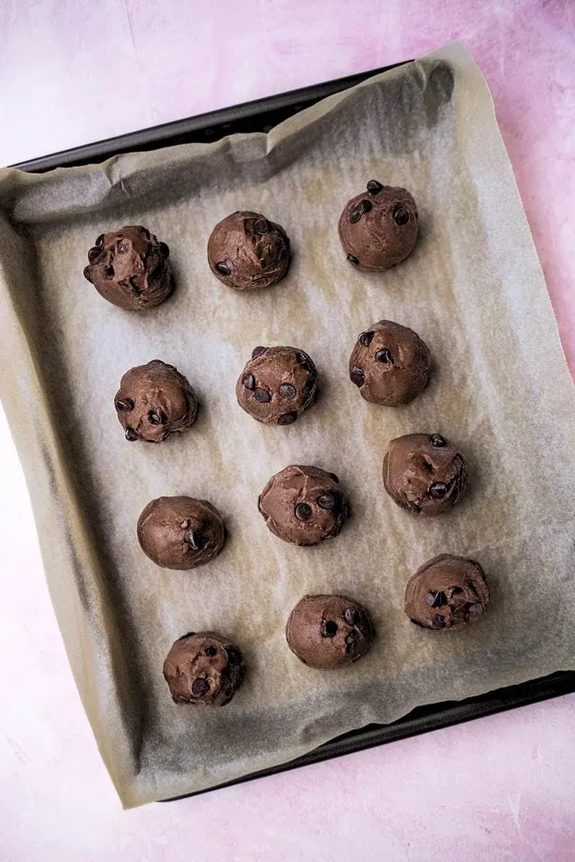 A top-down view of a baking sheet lined with parchment paper, filled with twelve balls of chocolate cookie dough.