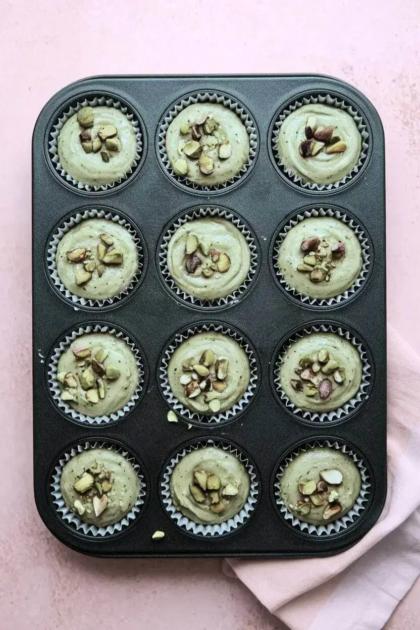 A top-down view of a muffin tin lined with paper, filled with pistachio muffin batter and topped with chopped pistachios.