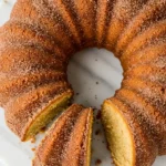 Apple cider donut cake with a slice removed, placed on a white marble plate against a white marble surface.