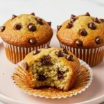 Banana chocolate chip muffins on a light pink plate with one muffin having a bite taken out of it.
