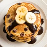 Banana Chocolate Chip Pancakes on a white plate drizzled with chocolate syrup.