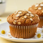Banana oatmeal muffin on a white plate and some marguerites are scattered around the plate.