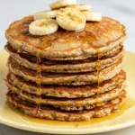 Banana oatmeal pancakes on a light-yellow plate topped with banana slices and some honey.