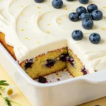 Blueberry lemon cake on a white baking pan against a light-yellow surface with a slice missing.