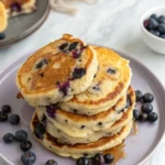 Blueberry pancakes on a light purple plate with some fresh blueberries around.