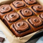 Chocolate cinnamon rolls in a lined baking pan topped with chocolate frosting.