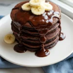 Chocolate pancakes placed on a white plate, drizzled with chocolate sauce and topped with banana slices.