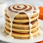 A stack of cinnamon roll pancakes on a white plate and topped with a cream cheese glaze.
