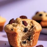 Gabs raisin muffin set on a light purple surface, with a bite taken out of it.