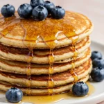 Greek yogurt pancakes on a white plate against a light pink surface.