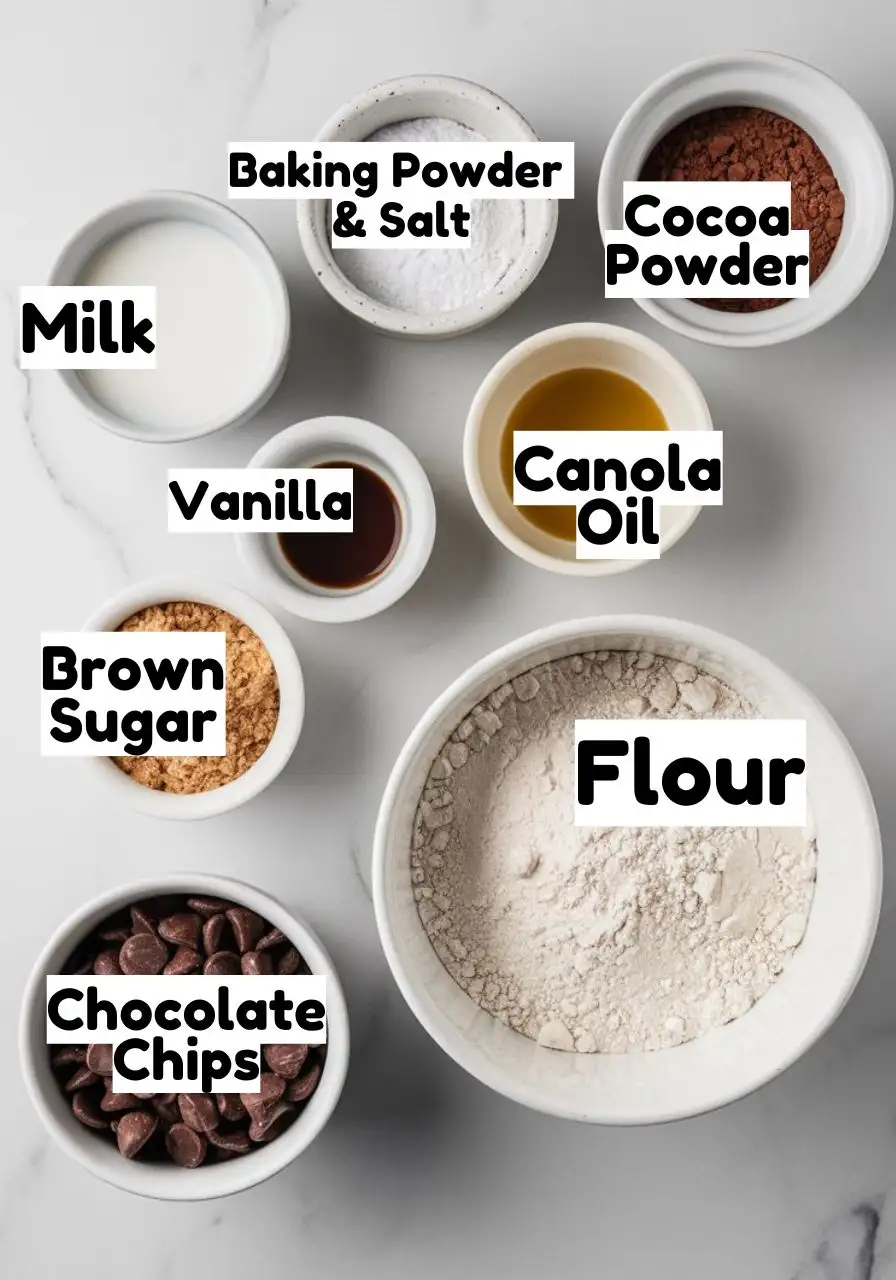 Ingredients on a white marble surface including flour, salt, baking powder, cocoa powder, vanilla, brown sugar, milk, and chocolate chips.