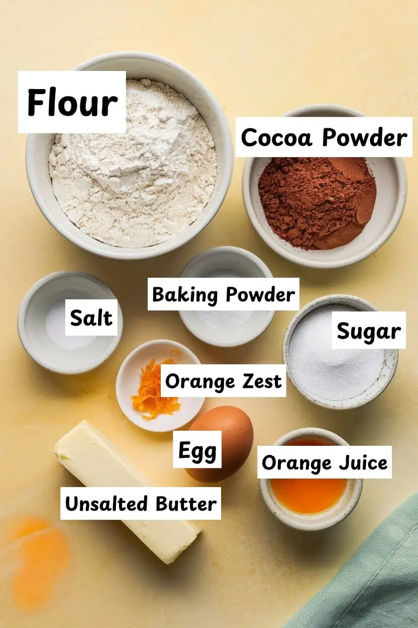 Ingredients on a light yellow surface including flour, cocoa powder, sugar, baking powder ,orange zest, orange juice, butter, and one egg.