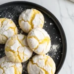 Lemon crinkle cookies on a black plate against a white marble surface.