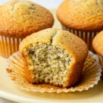 Lemon poppyseed muffins on a light-yellow plate with one muffin has a bite taken out of it.