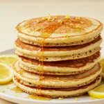 Lemon ricotta pancakes on a white plate drizzled with maple syrup and topped with a tiny lemon zest.