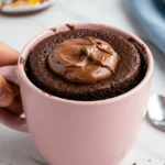 Nutella mug cake in a pink mug against a white marble surface and a blue napkin next to it.