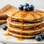 Stack of oat flour pancakes on a white plate topped with maple syrup and blueberries.
