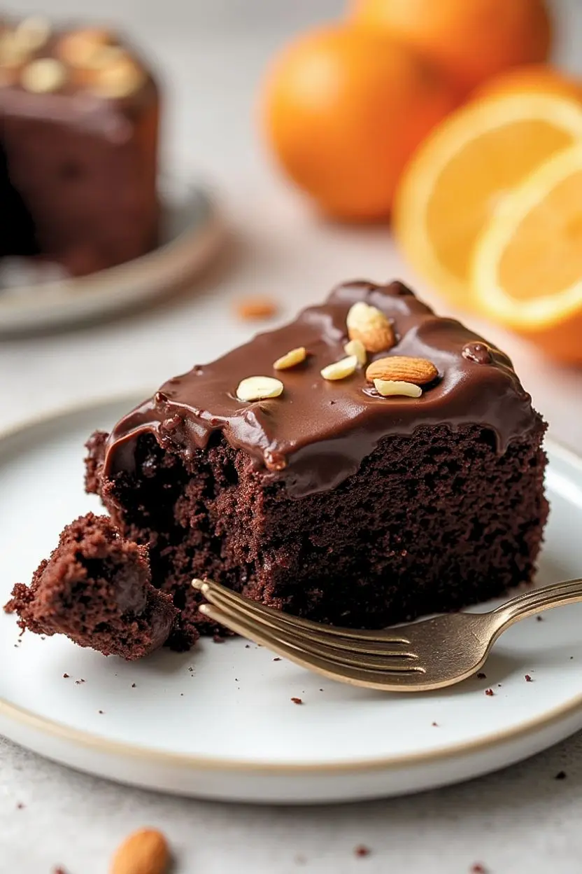 A slice of Prolific oven chocolate orange almond cake on a white plate and a golden fork next to it.