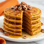 Pumpkin pancakes with a serving taken out of it on a white plate against a white marble surface.