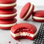 Red velvet whoopie pies on a gray surface and one of them with a bite missing.