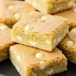 White chocolate brownies on a black plate against a white marble surface.