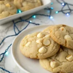 White chocolate macadamia nut cookies on a white plate against a white marble surface and blue LED strings around the plate.
