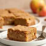 An apple brownie placed on a white plate with a fork next to it.