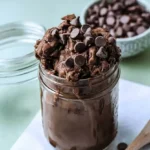 Edible chocolate batter in a glass jar topped with chocolate chips, with a wooden spoon beside it and a bowl filled with chocolate chips behind.