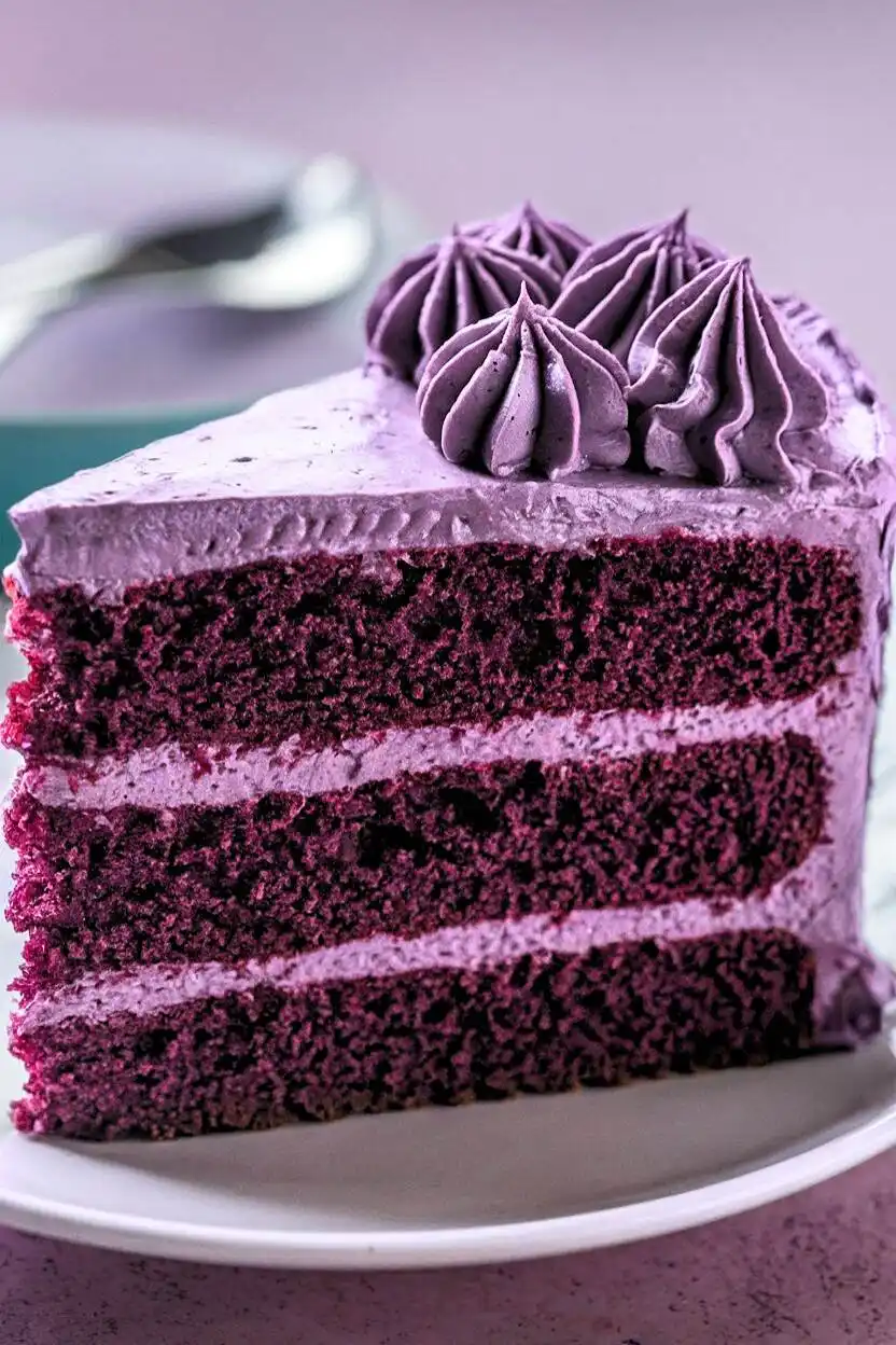 Slice of purple velvet cake placed on a white plate against a light purple surface.