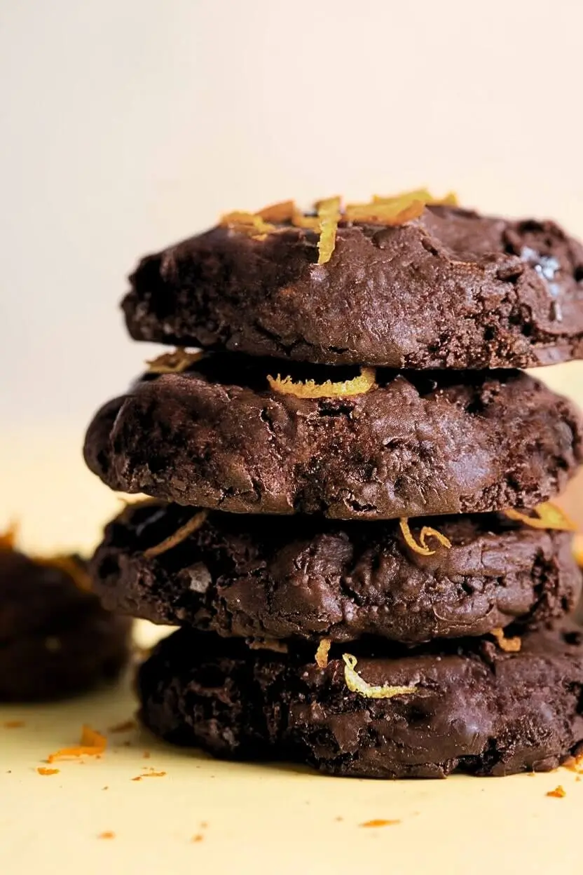 stack of orange chocolate cookies serbian sits on a light yellow surface garnished with orange zest.
